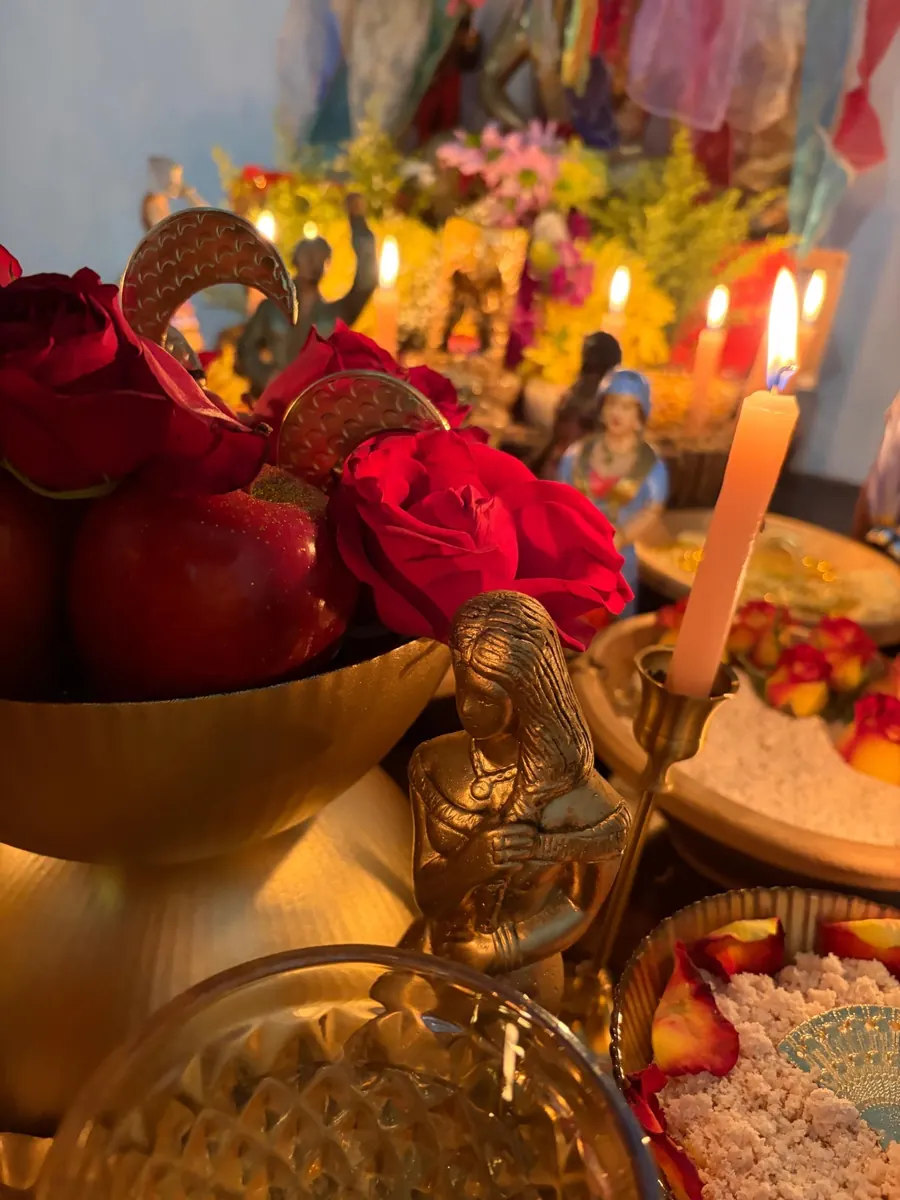Rosas vermelhas e maçãs em uma taça dourada ao lado de uma estatueta, com velas ao fundo